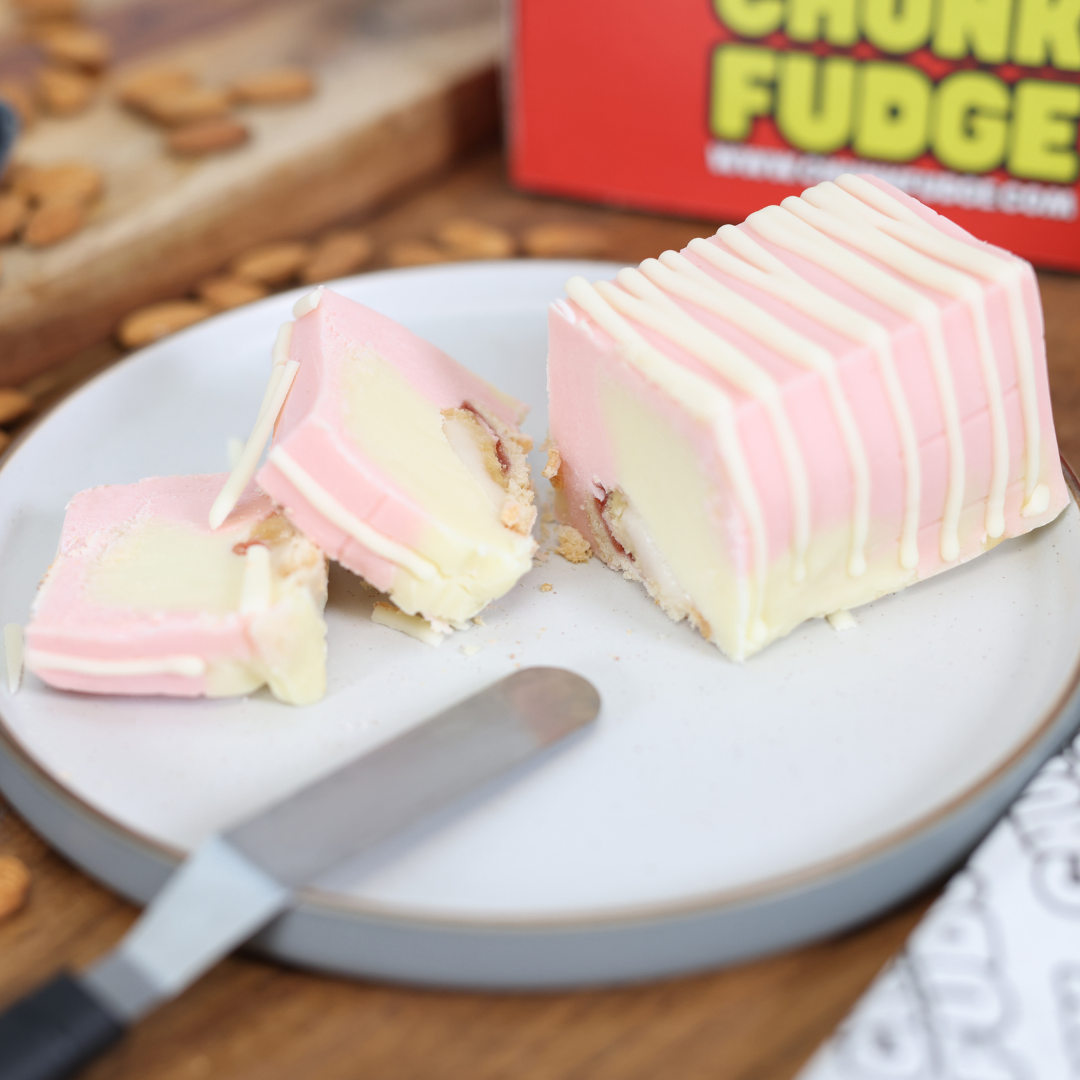 A slice of pink and white layered Bakewell Tart Fudge with a crunchy base sits on a plate, partially cut with a knife. In the background, a red CHUNK FUDGE box and scattered pretzels complete this tempting scene.