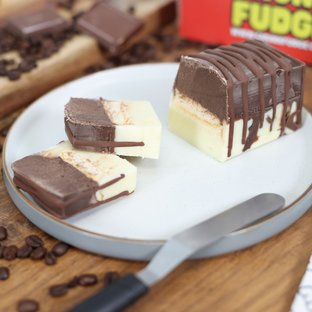 A plate holds slices of Tiramisu Fudge—rich chocolate and vanilla layers with chocolate drizzle. A butter knife rests nearby, with coffee beans scattered on the wooden table.
