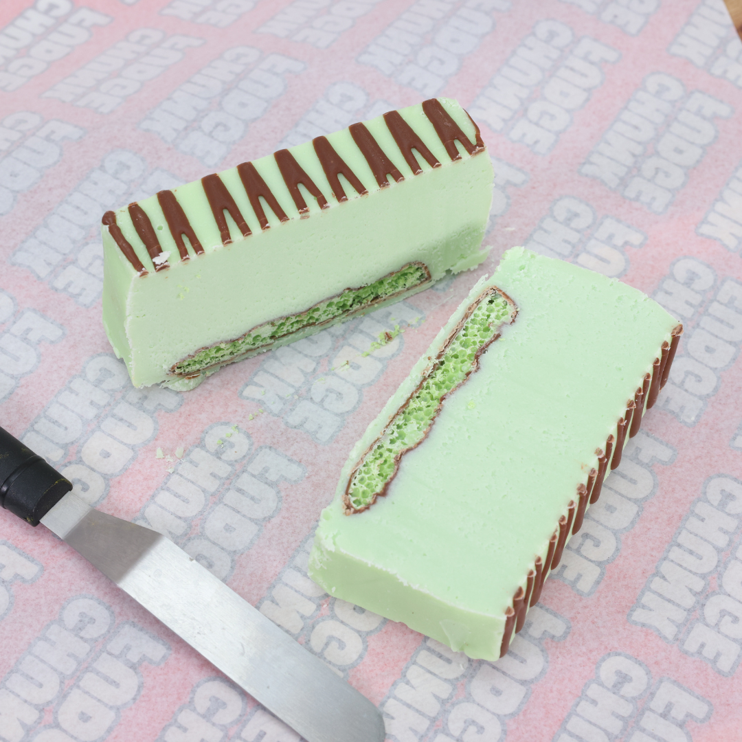 A rectangular Mint Aero Fudge slice with a chocolate drizzle, cut to reveal a chocolate cookie layer, sits on branded paper beside a metal spatula—perfect for fans of mint and chocolate treats.