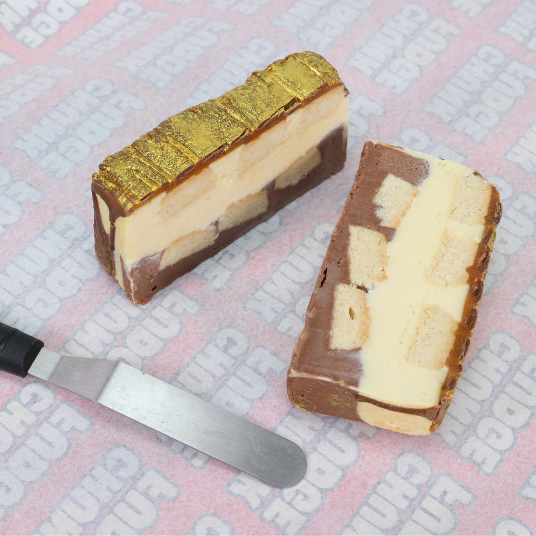 Two slices of Billionaires Fudge ice cream cake—layers of chocolate, vanilla, and fudge, one with a gold topping—are served on pink END CLINE branded paper beside a knife.