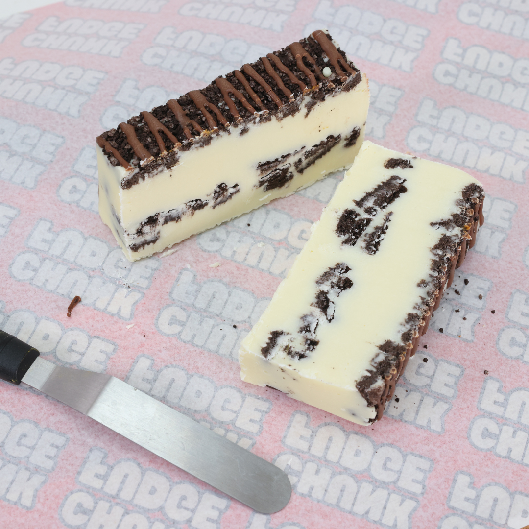 Two rectangular slices of Cookies And Cream Fudge rest on pink paper. Beside them is a cake spatula. The dessert features white ice cream, fudge drizzle, and crunchy Oreo cookie pieces in layered chocolate cookie crusts.